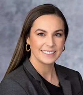 Headshot of Kelsey Gislason. She is a Caucasian woman with long brown hair. She is wearing a black shirt with a dark grey blazer on top and gold hoop earrings